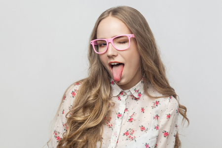 Playfully young woman in pink eyeglasses, looking at camera, tongue out and flirt with you. Studio shot. Isolated on gray background
の写真素材