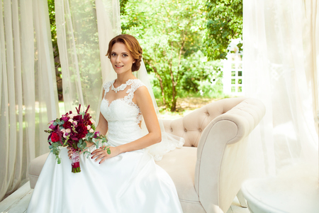 attractive bride with bouquet of flower wearing wedding white dress sitting on beige sofa in parkの写真素材