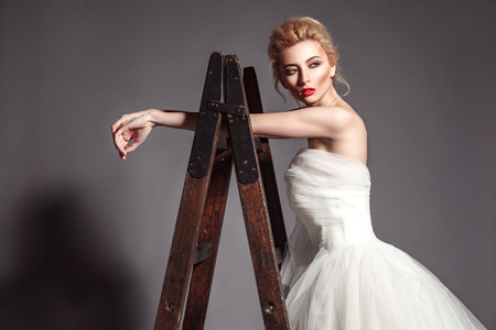 portrait in vogue style of fashion beautiful bride in wedding dress leaning on wooden ladder on grey background.の写真素材