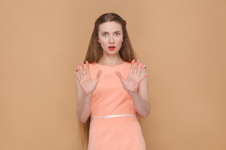 shocked face of panic woman, looking at camera. portrait of emotional cute, beautiful woman with makeup and long hair in pink dress. indoor, studio shot, isolated on light brown or beige background.の写真素材