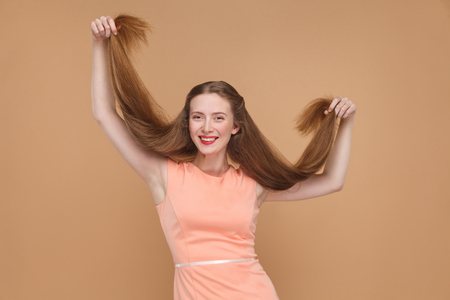 funny girl holding her long hairs and looking at camera with toothy smile. portrait of emotional cute, woman with makeup and in pink dress. studio shot, isolated on light brown or beige background.の写真素材