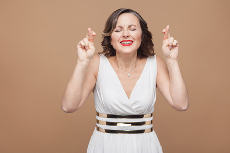hopeful middle aged woman. Crossed hands and closed eyes. Emotional expressing woman in white dress, red lips and dark curly hairstyle. Studio shot, indoor, isolated on beige or light brown backgroundの写真素材
