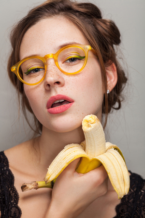 portrait of seductive woman in glasses with opened mouth holding banana and looking at camera. indoor, studio shot on gray background.の写真素材