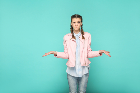 confused doubtful portrait of beautiful cute girl standing with makeup and brown pigtail hairstyle in striped light blue shirt pink jacket. indoor, studio shot isolated on blue or green background.の写真素材