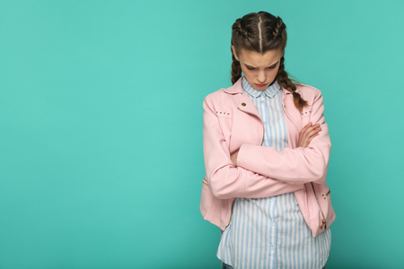 sad upset or depressed portrait of beautiful cute girl standing with makeup and brown pigtail hairstyle in striped blue shirt pink jacket. indoor, studio shot isolated on blue or green background.の写真素材