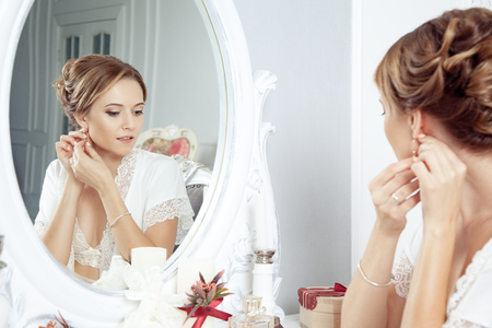 Young happy woman sitting in front of mirror and preparing to wedding. holding and wearing earrings in front of mirror. amazing bridal morning. wedding concept and feelings. indoor studio shot.の写真素材