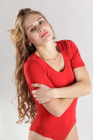 beautiful sexy girl in red bodysuit with long curly blonde hairstyle and makeup and perfect body standing with crossed hands and looking at camera. indoor studio shot on gray background.の写真素材