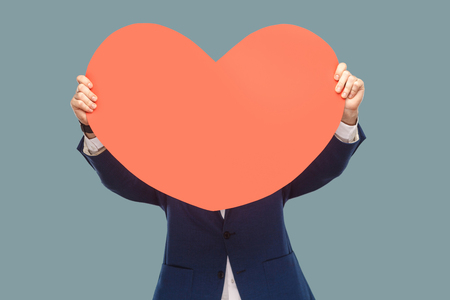Handsome businessman in blue jacket standing and holding and covering red big heart shape in front of face for copy space. Indoor, studio shot isolated on light blue background.の写真素材