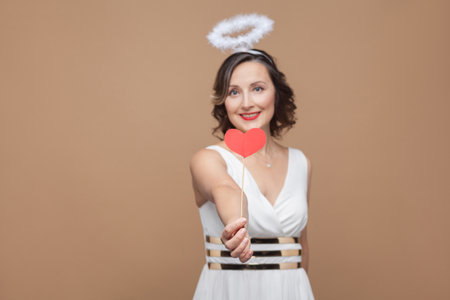 Middle aged angel brunette woman in white dress with nimbus on top of her head giving red heart sticker and looking at camera with toothy smile. Studio shot, indoor, isolated on light brown backgroundの写真素材