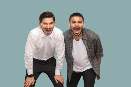 Portrait of two handsome bearded happy positive friends or partners standing and looking at camera and laughing with open mouth. indoor studio shot, isolated on light blue background.の写真素材