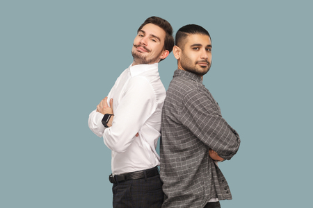 portrait of two handsome bearded successful friends or partners standing with crossed arms, looking at camera with proud face. profile, side view. indoor studio shot, isolated on light blue backgroundの写真素材