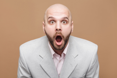are you serious? Portrait of handsome middle aged bald bearded businessman in classic light gray suit standing looking at camera with amazed face. indoor studio shot, isolated on brown background.の写真素材