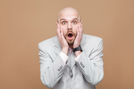 wow, its unbelievable. Portrait of amazed handsome middle aged businessman in light gray suit standing, holding hands on face and looking at camera with shocked face. studio shot on brown background.の写真素材