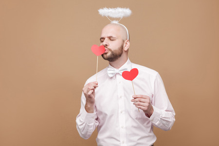 Happy handsome middle aged bald bearded angel in shirt and white halo on head standing and holding two red heart stickers and kissing one of them. studio shot, isolated on light brown background.の写真素材