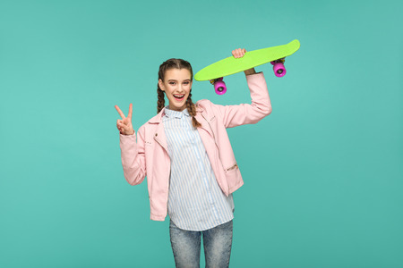 funny beautiful girl in casual style, pigtail hair and pink jacket, standing and holding skateboard and looking at camera with peace or victory sign. isolated, studio shot on blue or green backgroundの写真素材