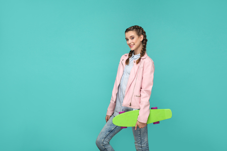happy beautiful girl in casual style, pigtail hair and pink jacket, standing and holding skateboard and looking at camera with toothy smile, Indoor isolated, studio shot on blue or green backgroundの写真素材