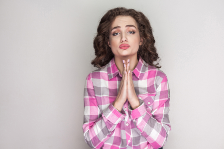 Pleased beautiful girl with pink checkered shirt, curly hairstyle and makeup standing looking at camera and requesting to forgive or helping. indoor studio shot, isolated on gray background.の写真素材