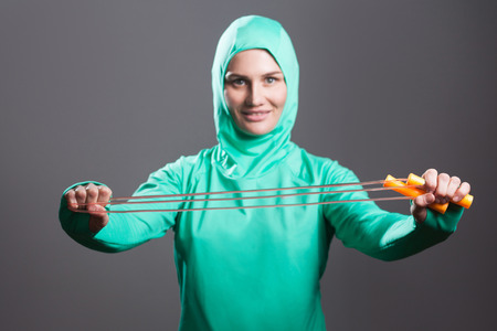 Happy beautiful muslim woman in green hijab or islamic sport wear standing, holding and showing jump rope and looking at camera with toothy smile. indoor studio shot, isolated on dark grey background.の写真素材