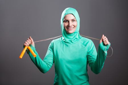Happy beautiful muslim woman in green hijab or islamic sport wear standing, holding jump rope and looking at camera with toothy smile. indoor studio shot, isolated on dark grey background.の写真素材