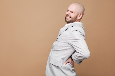 Back spine pain. Profile side view portrait of middle aged bald bearded businessman in light gray suit standing and touching his painful spin. indoor studio shot, isolated on light brown background.の写真素材