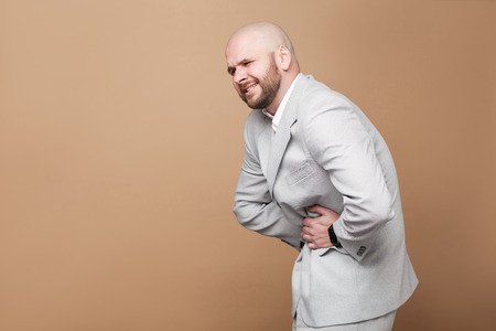 Stomach pain. profile side view portrait of middle aged bald bearded businessman in light gray suit standing and holding his painful belly. indoor studio shot, isolated on light brown background.の写真素材