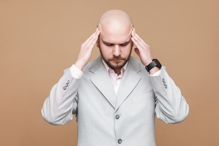 Headache and pain. Portrait of middle aged bald bearded businessman in light gray suit standing with painful head. indoor studio shot, isolated on light brown background.の写真素材