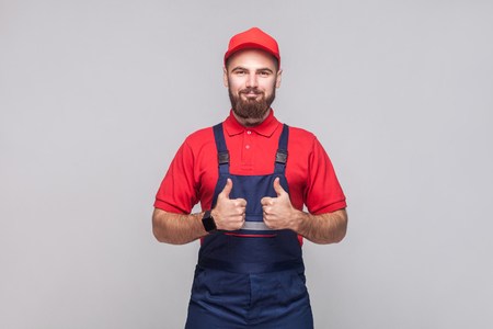 Work are done! Portrait of young satisfied cheerful repairman with beard in blue overall, red t-shirt and cap, standing and showing thumps up with smile. Grey background, indoor studio shot isolated.の写真素材