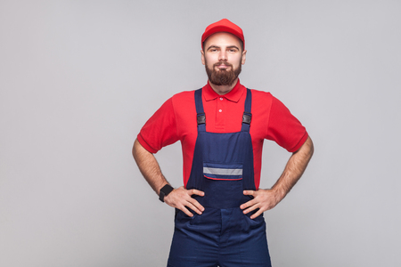 Portrait of young confident repairman with beard in blue overall, red t-shirt and cap standing and holding hands on waist with smile, indoor, studio shot, isolated on gray background, copy space.の写真素材