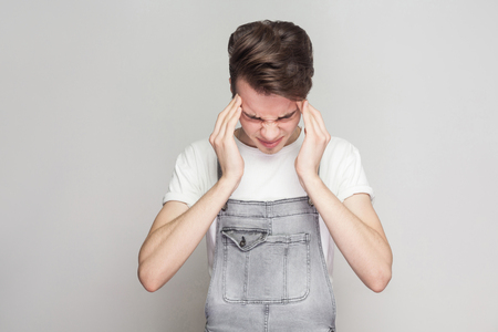 Portrait of young man suffering from severe headache, pressing fingers to temples, closing eyes to relieve pain with helpless face expression. Isolated, studio shot, copy space, on grey backgroundの写真素材