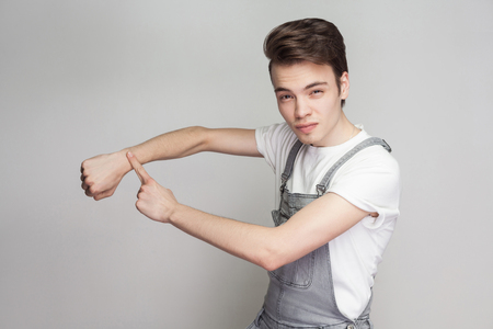 Serious young brunette man in casual style with white t-shirt and denim overalls standing, looking at camera and showing time gesture on hand. indoor studio shot, isolated on gray background.の写真素材