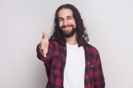 Happy smiley handsome man with beard and black long curly hair in casual style, checkered red shirt standing giving handshake and greeting with smile. indoor studio shot, isolated on grey background.の写真素材