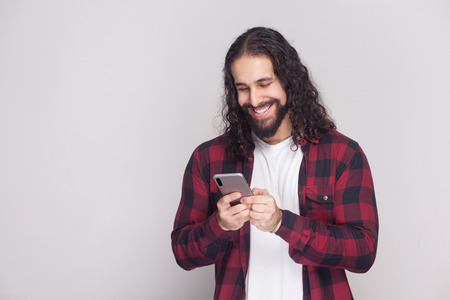 Cheerful handsome young man in red checkered shirt and long curly hair standing, using smartphone and texting message with toothy smile. Indoor, studio shot, isolated on grey background.の写真素材