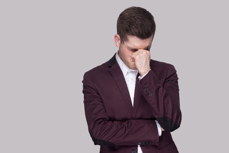 Portrait of sad depressed handsome young man in violet suit and white shirt, standing holding head down and hands on face and crying. indoor studio shot, isolated on grey background.の写真素材