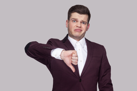 Portrait of handsome confused young man in violet suit and white shirt, standing, looking at camera with thumbs down and unsatisfied face. indoor studio shot, isolated on grey background.の写真素材