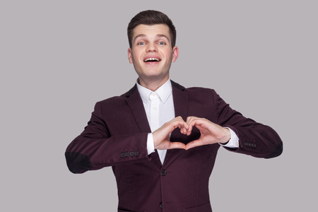 Portrait of happy funny handsome young man in violet suit and white shirt, standing, looking at camera with heart gesture sign. indoor studio shot, isolated on grey background.の写真素材
