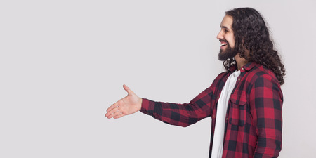 Profile side view of happy handsome man with beard and black long curly hair in checkered shirt standing, greeting with toothy smile and handshake. indoor studio shot, isolated on grey background.の写真素材