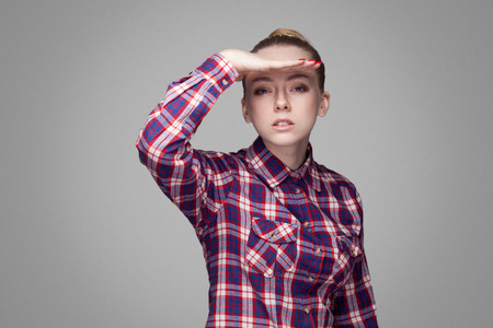 portrait of amazed beautiful blonde girl in pink checkered shirt, collected bun hairstyle and standing and looking at camera with hand on head and open mouth. studio shot, isolated on gray backgroundの写真素材