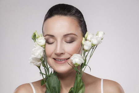 Closeup portrait of cheerful beautiful woman with perfect skin and aged wrinkles are lean on flowers. Skin Care Concept, aging cosmetics, isolated on grey background, copy space, studio shot, indoorの写真素材
