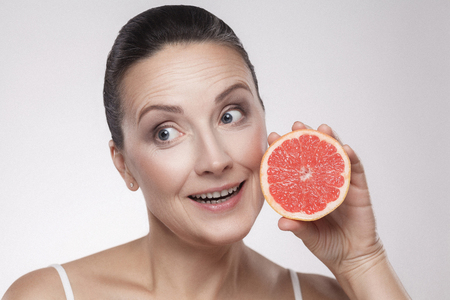 Closeup portrait of charming, pretty, attractive woman with perfect skin smiling after cream, balm, mask, lotion, holding half of grapefruit isolated on grey background. Indoor,studio shot,copy spaceの写真素材
