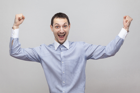 Portrait of excited joyful handsome bristle businessman in classic light blue shirt standing with raised arms and celebrating his victory. indoor studio shot, isolated on grey background copyspace.の写真素材