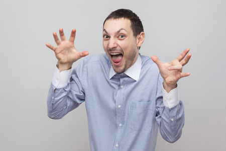 Portrait of amazed funny handsome bristle businessman in classic light blue shirt standing with raised hands and crazy face, screaming. indoor studio shot, isolated on grey background copyspace.の写真素材