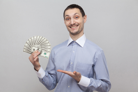 Portrait of satisfied attractive young adult businessman in blue shirt standing, giving and pointing with finger fan of money and toothy smile. Indoor studio shot,isolated, gray background, copy spaceの写真素材