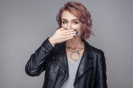 Portrait of shocked beautiful girl with short hairstyle, makeup in casual style black leather jacket standing, looking at camera and covering her mouth. indoor studio shot, isolated on grey backgroundの写真素材