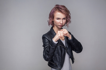 Portrait of serious beautiful girl with short hairstyle and makeup in casual style black leather jacket standing with boxing fist and looking at camera. indoor studio shot, isolated on grey backgroundの写真素材