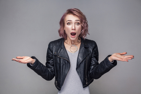 Portrait of shocked girl with short hairstyle, makeup in black leather jacket standing raised arms and looking at camera with big eyes and open mouth. indoor studio shot, isolated on grey backgroundの写真素材