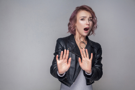 Please don't touch me. Portrait scared of beautiful girl with short hairstyle in casual style black leather jacket standing, blocking and looking. indoor studio shot, isolated on grey background.の写真素材