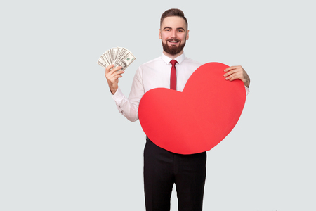 Young bearded man holding many dollars and red heart shape and looking at camera with toothy smile. Indoor, studio shot, isolated on gray backgroundの写真素材