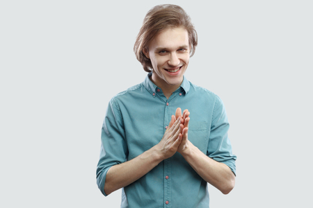 Portrait of cunning handsome long haired blonde young man in blue casual shirt standing and looking at camera with bitchy plan. indoor studio shot, isolated on light grey background.の写真素材