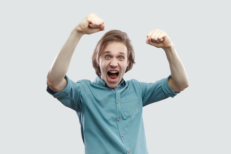 Portrait of amazed happy handsome long haired blonde young man in blue casual shirt standing screaming and celebrating his victory. indoor studio shot, isolated on light grey background.の写真素材