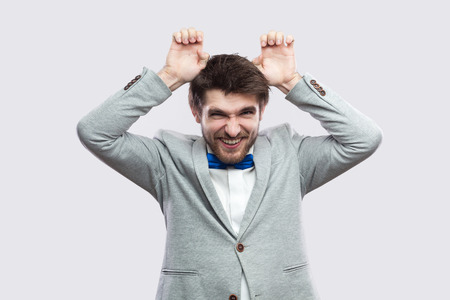 Portrait of funny handsome bearded man in casual grey suit and blue bow tie standing with comic face...